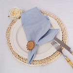 L'image montre une table de style rustique avec une assiette blanche, des couverts dorés et une serviette de table grise enroulée avec une épingle décorative en forme de fleur. Une rose blanche et un tapis de table en jonc sont également visibles, créant une composition élégante et chaleureuse.