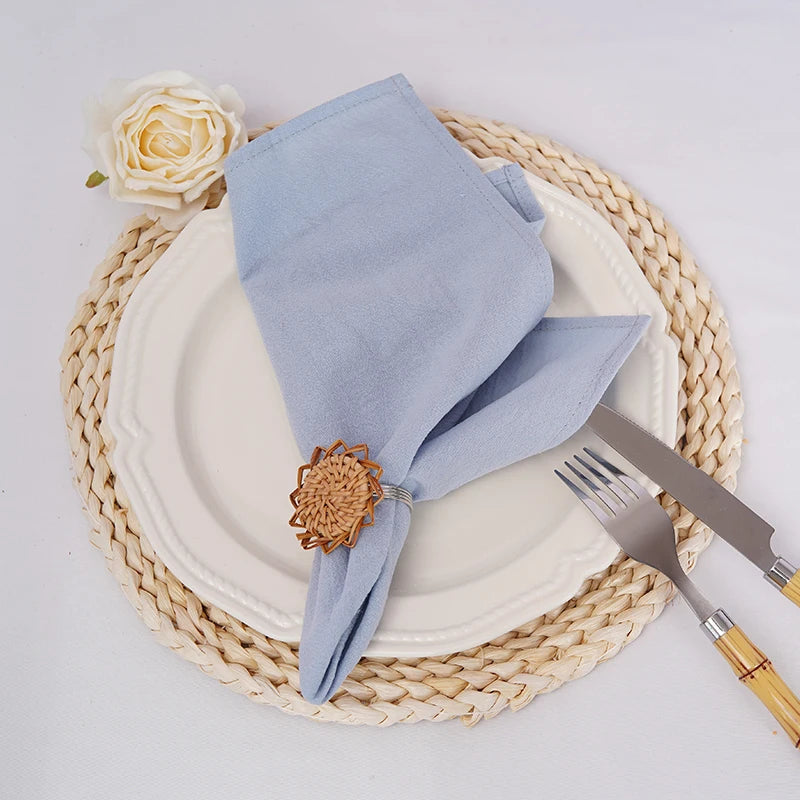 L'image montre une table de style rustique avec une assiette blanche, des couverts dorés et une serviette de table grise enroulée avec une épingle décorative en forme de fleur. Une rose blanche et un tapis de table en jonc sont également visibles, créant une composition élégante et chaleureuse.