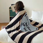 L'image montre un jeune individu enveloppé dans une couverture rayée avec un motif graphique et un texte, donnant une impression de confort et de détente.