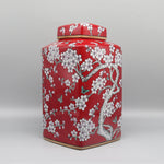 Un élégant pot de stockage en céramique de couleur rouge vif, décoré de magnifiques fleurs de prunier blanc et de papillons, évoquant une ambiance orientale chaleureuse et raffinée.