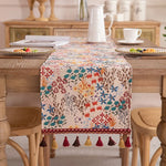 Cette image montre une table de salle à manger décorée avec un chemin de table coloré et orné de motifs floraux, avec des accessoires décoratifs comme des bougies et des tasses.