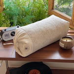 Sur une étagère en bois, on peut voir un rouleau de matériel textile beige avec un minuteur et une bougie dans un récipient en bois, le tout entouré de végétation verdoyante visible par la fenêtre.