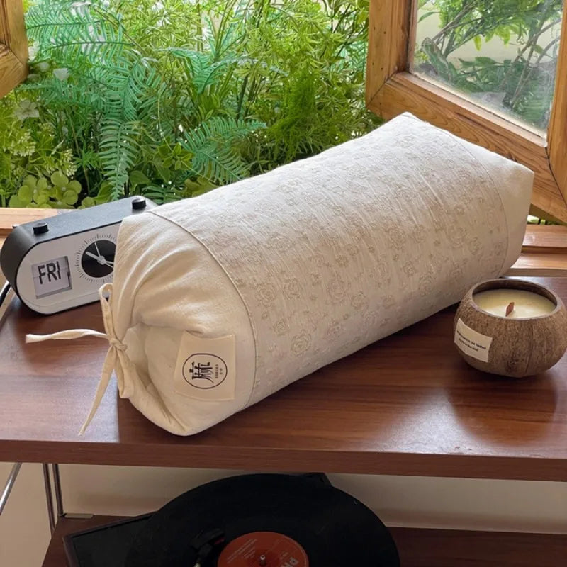 Sur une étagère en bois, on peut voir un rouleau de matériel textile beige avec un minuteur et une bougie dans un récipient en bois, le tout entouré de végétation verdoyante visible par la fenêtre.