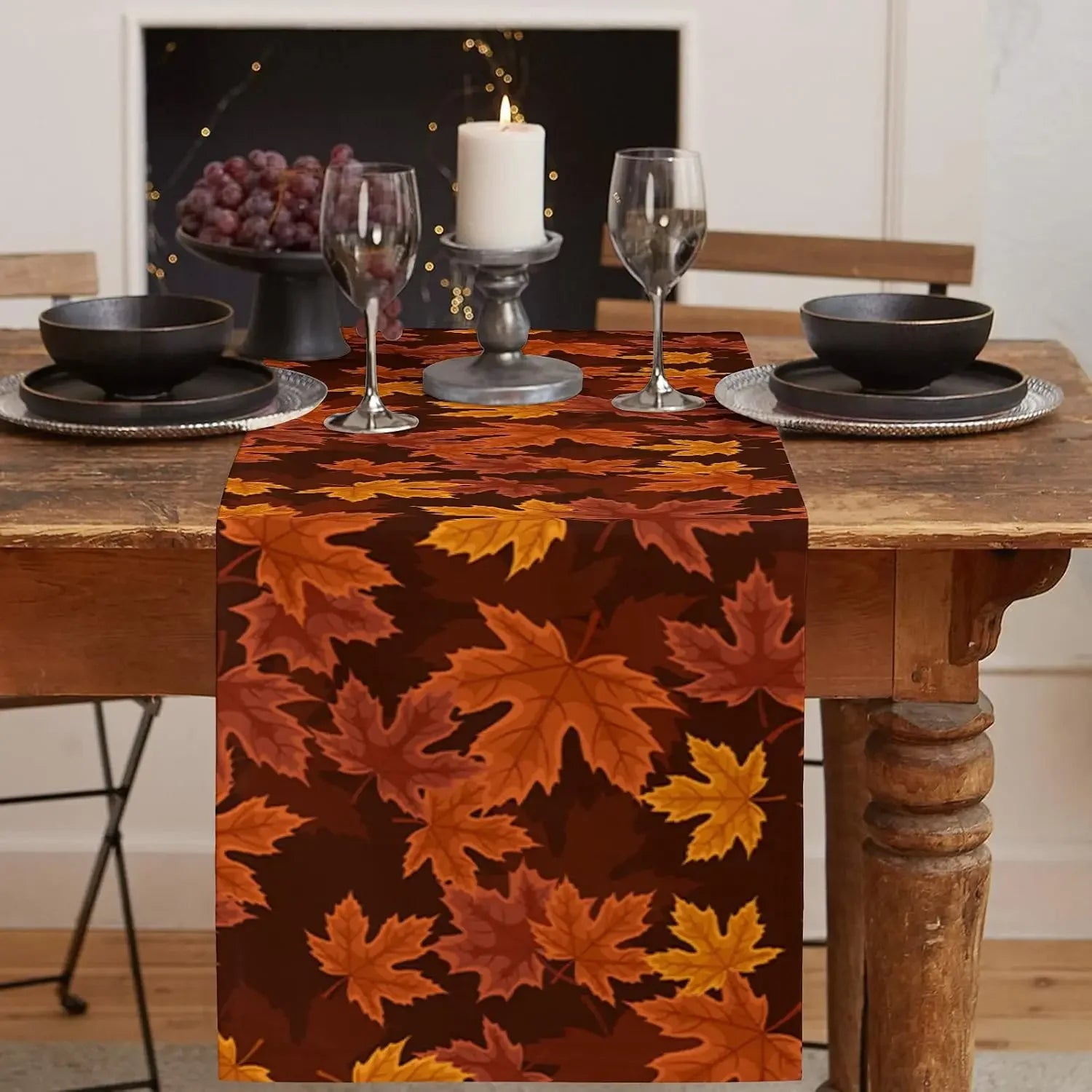 Une table de salle à manger avec des assiettes, des verres à vin et une bougie allumée, sur une nappe en velours imprimée de feuilles d'automne oranges et brunes.