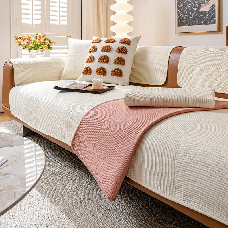 Un canapé blanc douillet avec des coussins décoratifs beiges et une couverture rose pastel, dans un salon lumineux avec des stores et des fleurs fraîches.