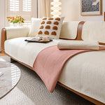 Un canapé blanc douillet avec des coussins décoratifs beiges et une couverture rose pastel, dans un salon lumineux avec des stores et des fleurs fraîches.