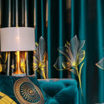 Lampe de table élégante avec un abat-jour blanc, posée près d'un canapé bleu richement décoré. En arrière-plan, des rideaux bleu foncé arborent des motifs floraux jaunes lumineux, créant une ambiance chaleureuse et moderne dans le salon. Accessoire idéal pour ajouter une touche de sophistication et de style à votre décoration intérieure.