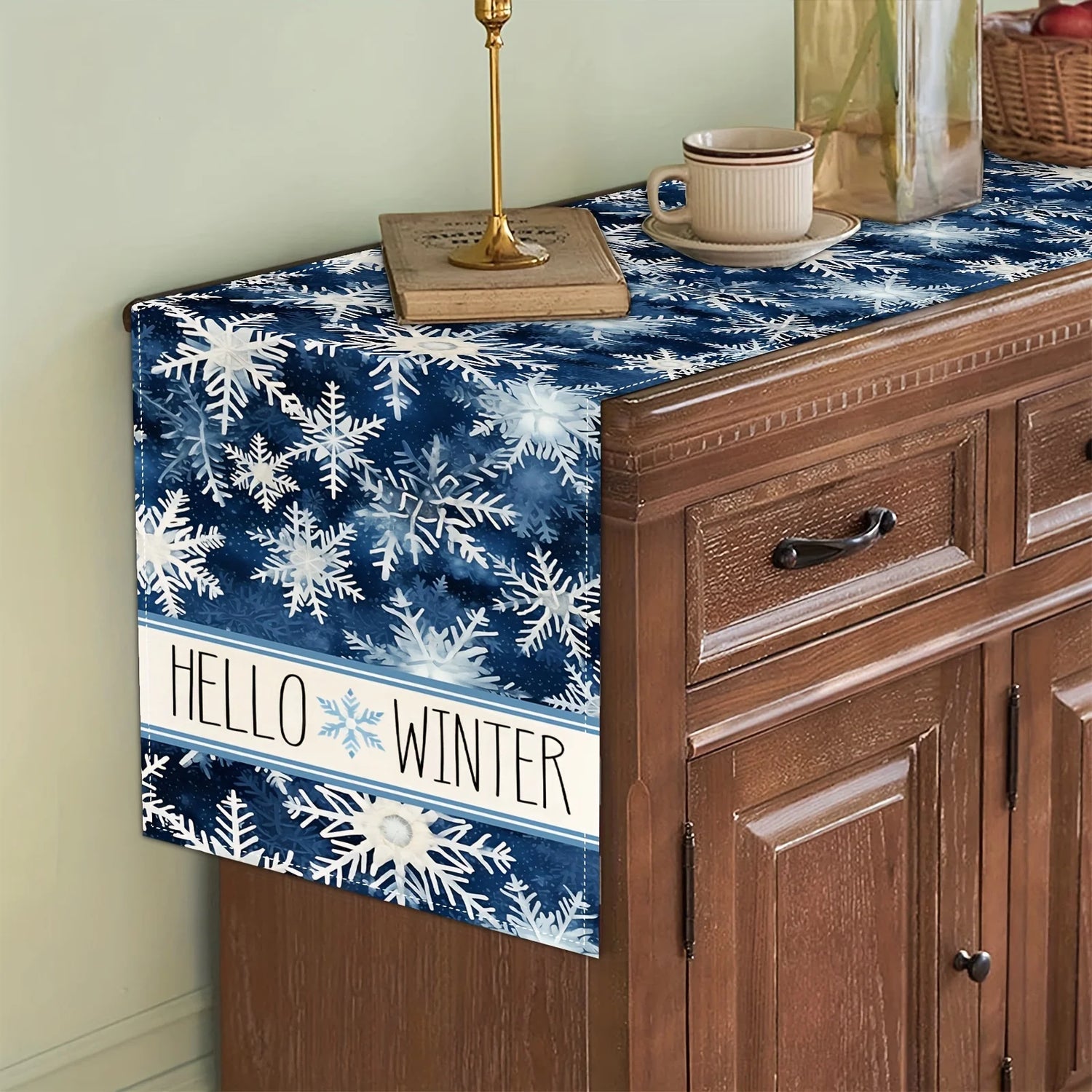 Une nappe de table bleu marine avec des motifs de flocons de neige et le texte "HELLO WINTER" au centre décore un meuble en bois rustique orné d'une lampe dorée et d'une tasse en céramique.