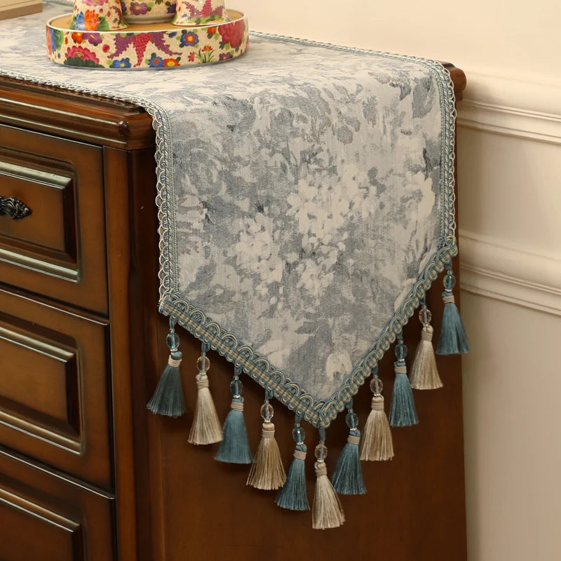 Un joli chemin de table orné de fleurs colorées et de glands de couleur beige et bleu turquoise, offrant une touche de style et d'élégance à ce meuble de bois.