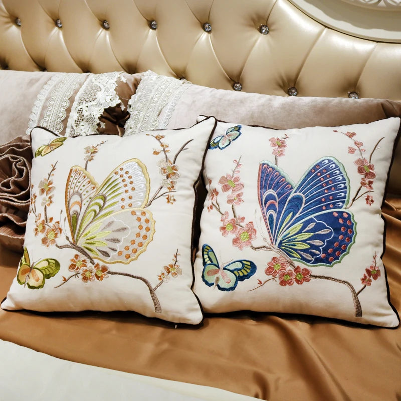 Deux oreillers blancs brodés de magnifiques papillons et de fleurs colorées, reposant sur un canapé à capitonnage doré, créant une atmosphère élégante et chaleureuse.