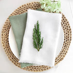 Une nappe en fibres tressées sert de support à des serviettes blanches avec une branche de conifère brodée, accompagnées de feuillages verts.