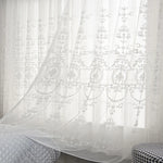Voilage blanc transparent à motifs floraux élégants, flottant légèrement devant une fenêtre dans une chambre paisible.