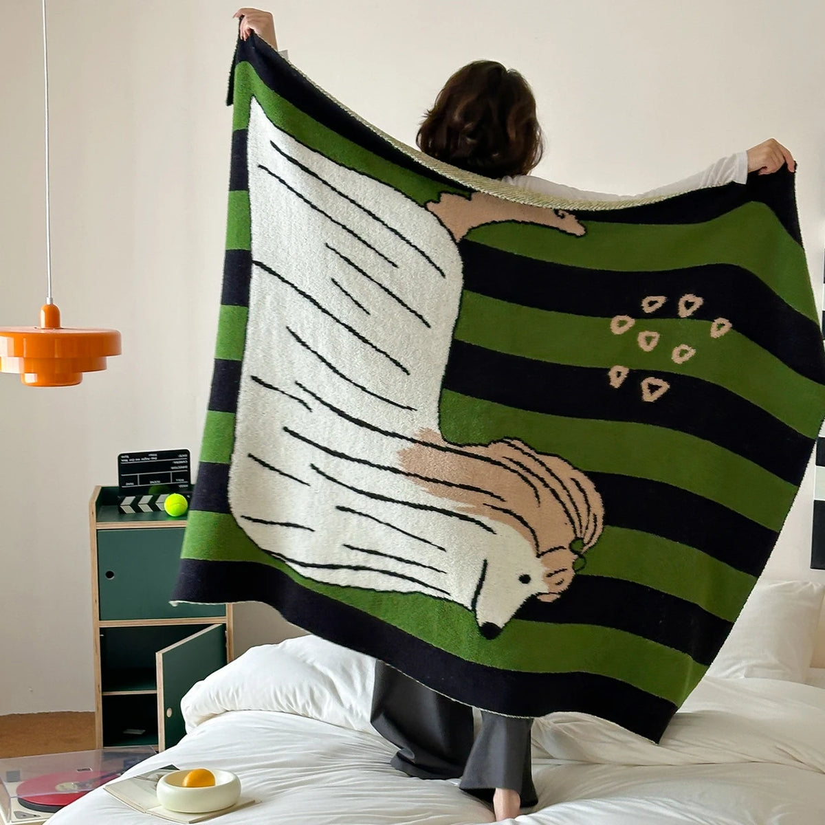 Une grande couverture à motif rayé vert et noir, avec un dessin de hérisson blanc, reposant sur un lit.