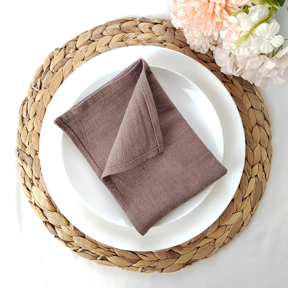 Une serviette de table marron pliée est placée sur une assiette blanche posée sur un napperon en fibres tressées autour de belles fleurs.