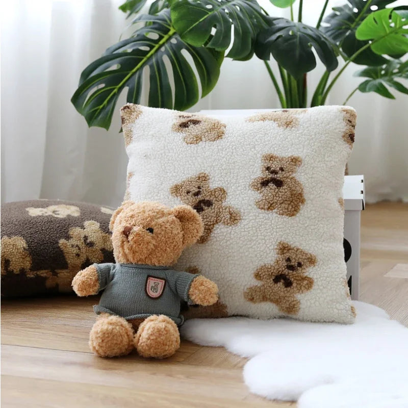 Un coussin décoratif moelleux avec un imprimé d'ourson et un nounours brun sur fond clair, placés près d'un pot de plantes vertes dans un intérieur chaleureux.