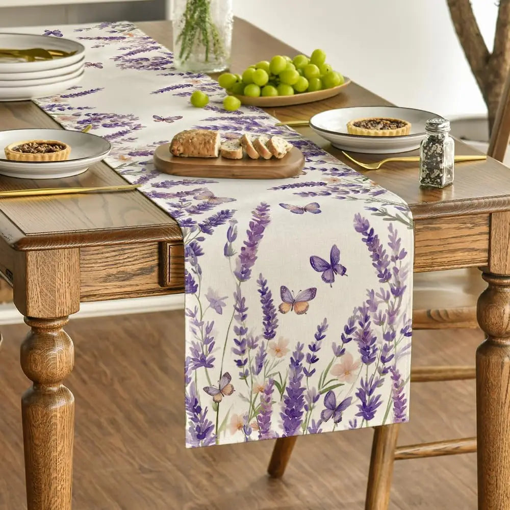 Nappe aux motifs floraux mauves avec des papillons, sur une table en bois avec des assiettes, un plat de raisins verts et un pain tranché.