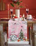 La table est magnifiquement décorée pour les fêtes, avec une nappe rose ornée de motifs hivernaux, un arbre de Noël décoré et une belle composition florale.