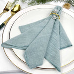 Une serviette en tissu bleu clair est élégamment posée sur une assiette blanche avec une fourchette et un couteau dorés.