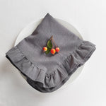 Sur un plat blanc, un linge de cuisine gris avec un motif de volants encadre trois cerises rouges fraîches avec leurs feuilles vertes, créant une scène élégante et raffinée.