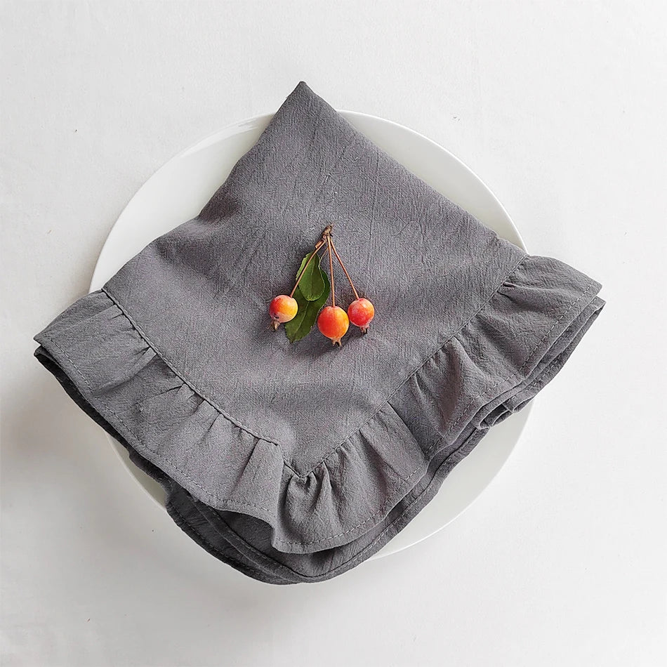 Sur un plat blanc, un linge de cuisine gris avec un motif de volants encadre trois cerises rouges fraîches avec leurs feuilles vertes, créant une scène élégante et raffinée.