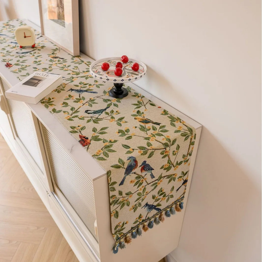Le meuble en bois blanc est recouvert d'un élégant chemin de table en tissu orné d'un motif floral coloré et de petits oiseaux, créant une ambiance chaleureuse et raffinée dans cette pièce.
