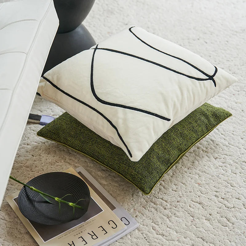 Coussin décoratif beige et vert avec motifs graphiques simples, posé sur un tapis, accompagné d'un magazine et d'un autre accessoire décoratif sombre.