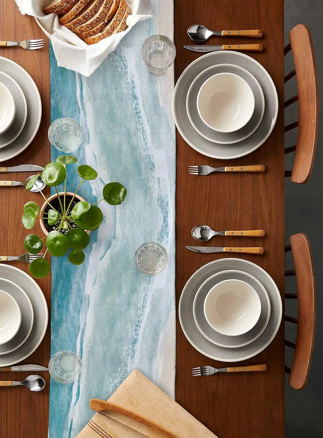 Cette image présente une table de salle à manger joliment dressée avec de la vaisselle en céramique grise, des ustensiles en bois et des éléments décoratifs tels que des verres et des plantes.