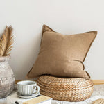 Une belle composition avec un coussin beige moelleux, un vase vintage et une tasse de thé posée sur un panier tressé, créant une atmosphère chaleureuse et cocooning.