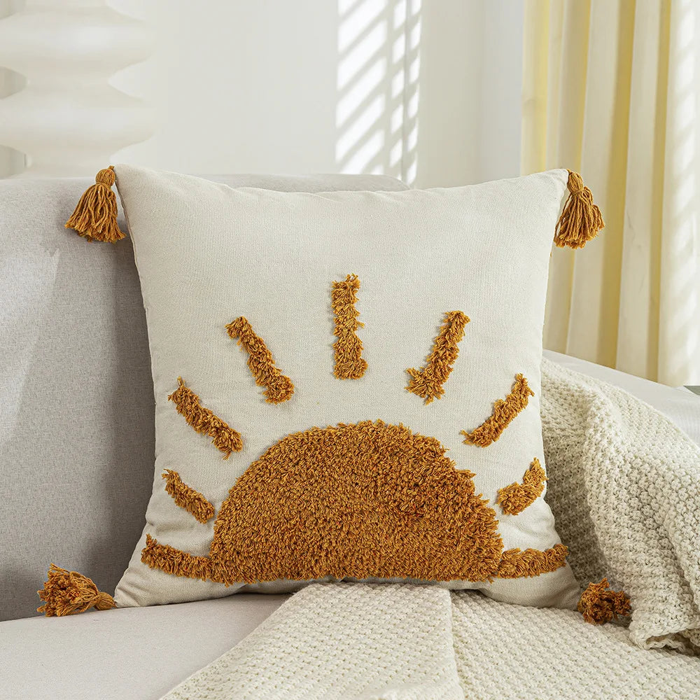 Coussin décoratif beige avec des motifs en peluche dorés évoquant le soleil sur une couverture blanche dans un intérieur lumineux.