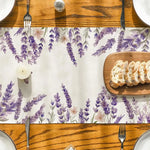 Cette image montre une table en bois avec des motifs de fleurs de lavande violettes sur un napperon, accompagnée de couverts en métal et de tranches de pain sur une planche en bois.