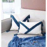 Coussin blanc et bleu avec franges, posé sur un canapé gris, accompagné d'une couverture en lin bleu.