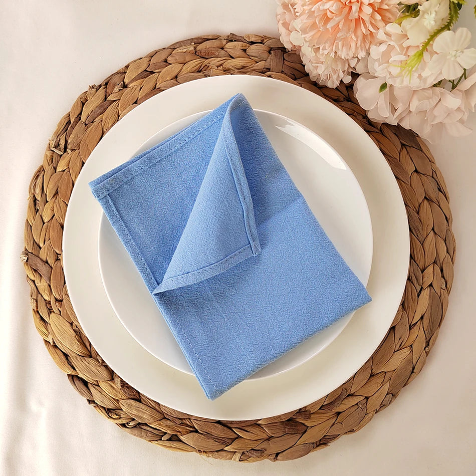 Assiette blanche avec une serviette de table bleu ciel pliée, posée sur un dessous de plat tressé en marron, entouré de fleurs roses.