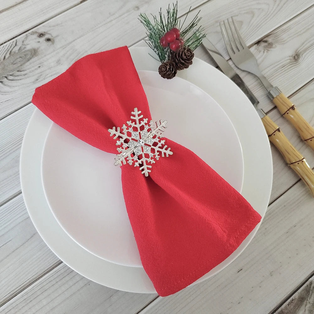 Une assiette blanche avec une serviette de table rouge portant un ornement en forme de flocon de neige, entouré de pommes de pin et d'épinettes vertes, sur une table en bois.