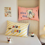 Deux oreillers colorés avec des motifs d'ours en peluche, l'un rose avec l'inscription "nice day" et l'autre jaune et blanc à rayures, complétés par une tasse et un petit gâteau, créant une atmosphère chaleureuse et accueillante.
