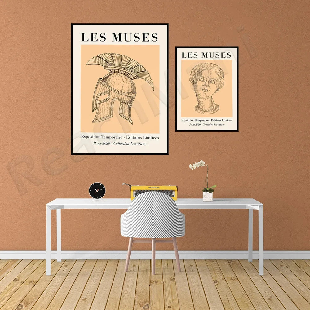 L'image présente des affiches d'exposition intitulées "Les muses", avec des illustrations d'un casque et d'un buste de statue grecque, sur un mur beige dans un espace de bureau avec une table, une chaise et quelques accessoires décoratifs.