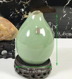 Un vase en forme de goutte d'eau de couleur vert pastel, posé sur un support en pierre noire avec des motifs pointillés, mesurant environ 9,6 cm de hauteur.