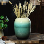 Un vase en céramique de couleur vert dégradé qui contient des épis de blé et autres plantes saisonnières, posé sur une table en bois.