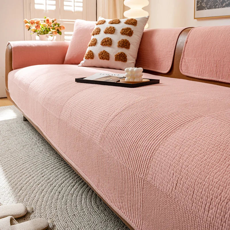 Un élégant canapé rose pâle avec des coussins décoratifs, placé dans un intérieur lumineux avec des fenêtres, un tapis gris et une belle fleur blanche sur une tablette noire.