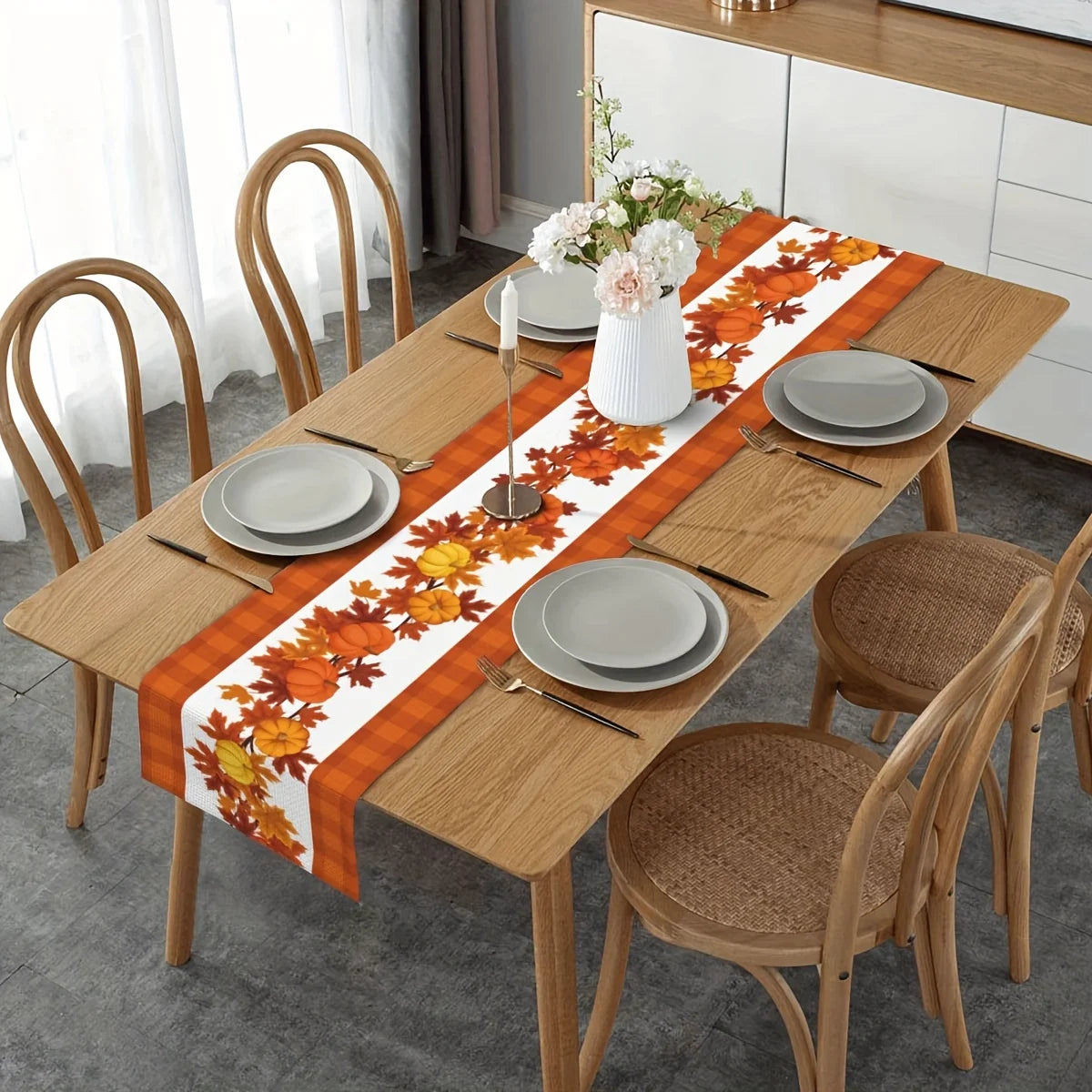 Une table de salle à manger en bois avec un chemin de table décoré de feuilles d'automne orange et de fleurs, entourée de chaises en bois assorties.