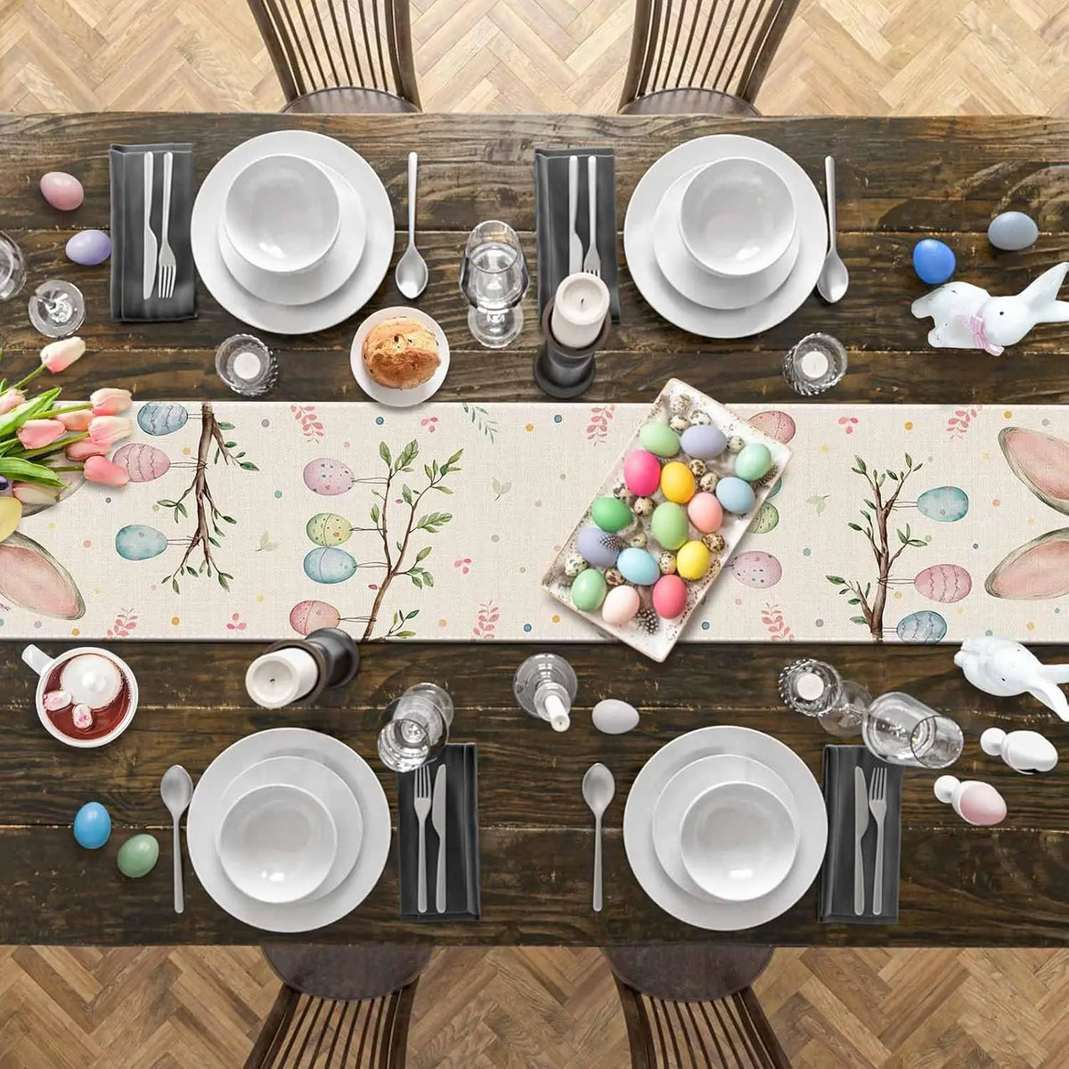 Table dressée pour Pâques avec une nappe ornée de motifs floraux et d'œufs colorés, entourée d'assiettes, de verres, de couverts et de décorations de Pâques.