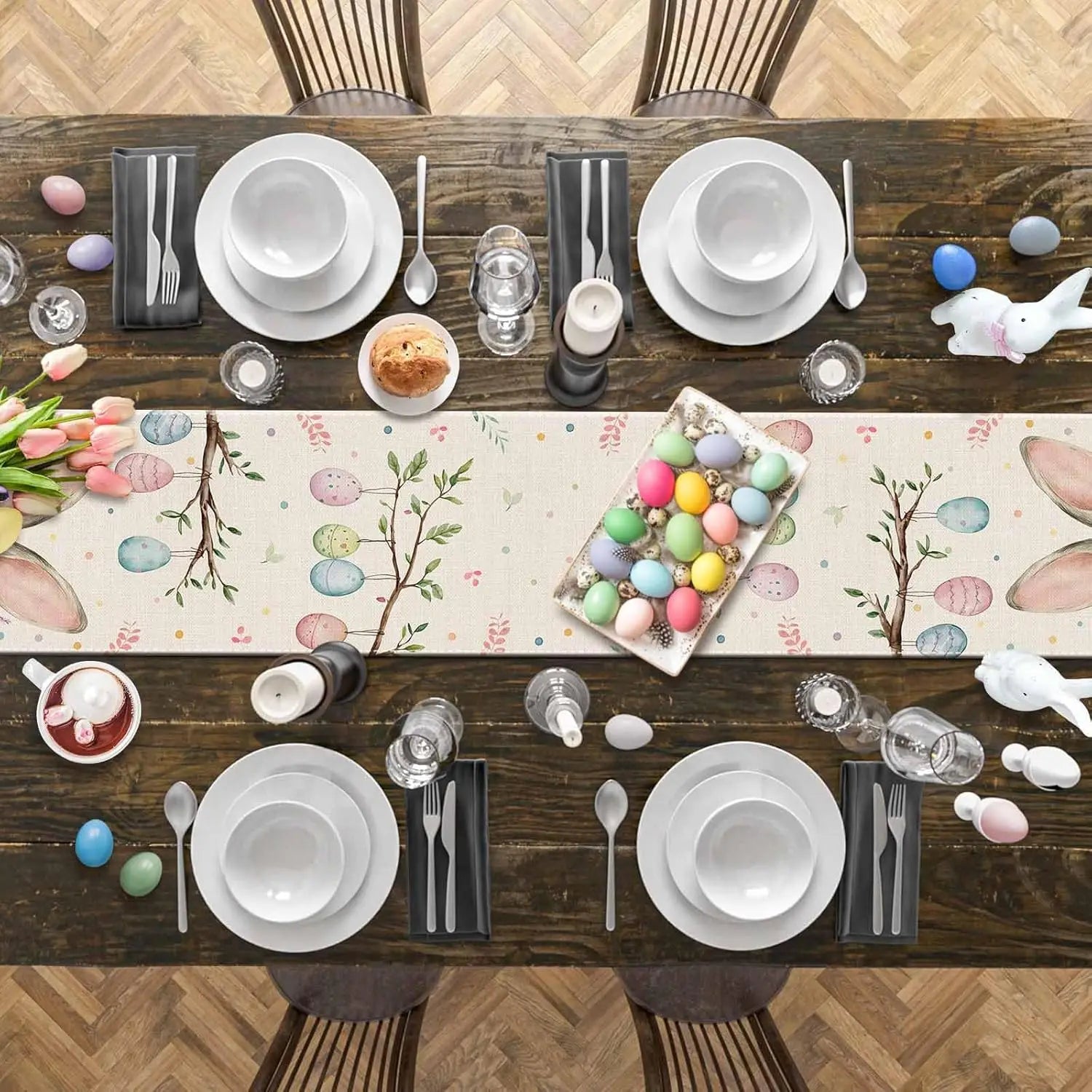 Table dressée pour Pâques avec une nappe ornée de motifs floraux et d'œufs colorés, entourée d'assiettes, de verres, de couverts et de décorations de Pâques.