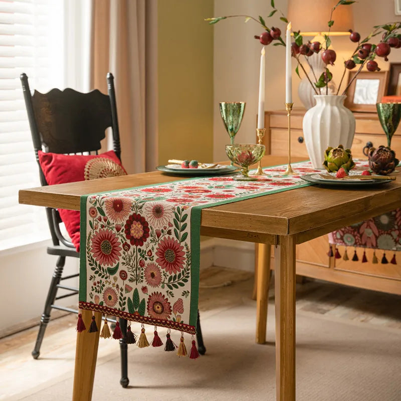La table en bois est recouverte d'un chemin de table coloré et orné de motifs floraux, entouré de chaises en bois foncé et décoré de bougies et de fleurs, créant une ambiance chaleureuse et conviviale.