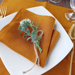 Une élégante serviette en lin ocre avec un bouquet de fleurs et de branches vertes attaché avec de la ficelle, posé sur une assiette blanche avec des couverts dorés.