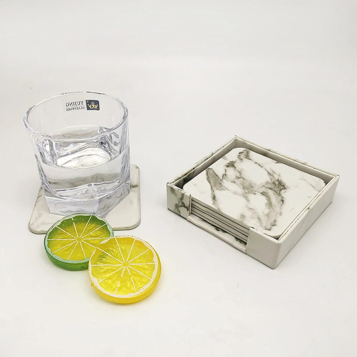 Un verre en cristal rempli d'eau et entouré de morceaux de citron vert et jaune, posé sur un support en marbre blanc avec une boîte de rangement en marbre.