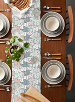 Une table joliment dressée avec des assiettes blanches, des couverts en bois et une nappe aux motifs colorés, mettant en valeur un arrangement végétal frais et harmonieux.