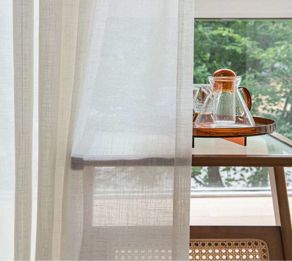 Gros plan sur un voilage blanc transparent devant une table en verre et un service à thé.