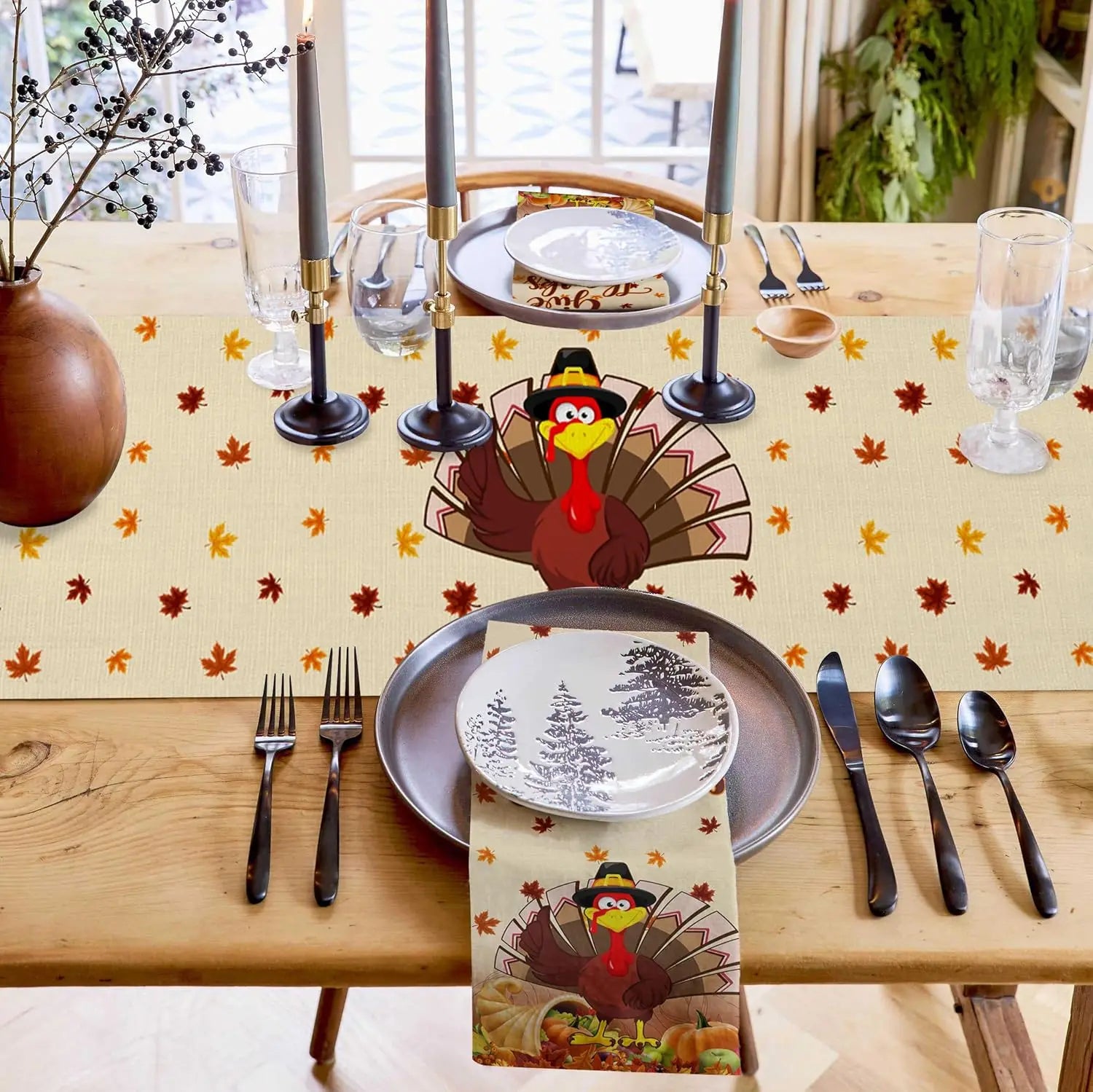 Table de fête d'automne avec un décor festif, assiettes avec motifs d'arbres enneigés, couverts noirs, bougies et une figurine de dindon avec chapeau de pèlerin sur le dessus.