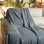 Une couverture décorative noire et grise avec un motif géométrique sur un canapé blanc, dans une pièce avec des plantes vertes.