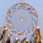 Cette image montre un magnifique capteur de rêves suspendu avec de belles perles colorées, des plumes et un pendentif brillant en son centre, créant une atmosphère apaisante et méditative.