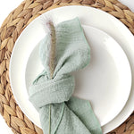Une serviette de table textile de couleur verte avec un motif texturé, posée sur une assiette blanche sur un napperon tressé.